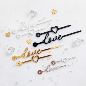 Puede incluir: Una variedad de palitos decorativos con la palabra "love" en escritura cursiva. Los palitos son blancos, negros, dorados y plateados, con detalles en forma de corazón. Pequeños corazones dorados y cristales transparentes están dispersos.