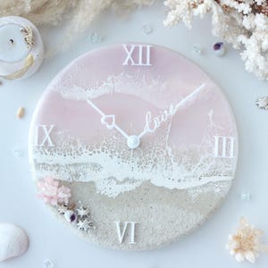 May include: A round wall clock featuring a resin design of a beach with light pink water, white foam waves, and a sandy shore. The clock face has white Roman numerals at the 12, 3, 6, and 9 positions, and white hands, one shaped like a heart.
