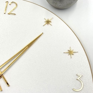 May include: Close-up of a white clock face with gold-colored hands and numerals. The clock features gold starburst markers at the 12, 3, and 6 o'clock positions. The clock's hands are positioned to show the time.