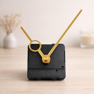 Puede incluir: Un mecanismo de reloj cuadrado negro con manecillas doradas y un cubo central. La manecilla de la hora es una barra corta y plana, y el minutero es una barra larga y delgada. Mecanismo para proyectos de relojería.