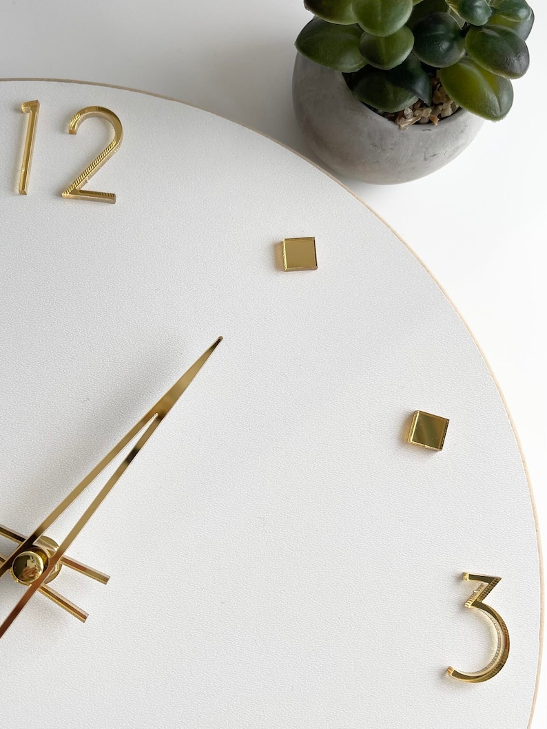 Peut inclure: Horloge murale blanche avec des aiguilles et des chiffres dor&eacute;s. Le cadran de l'horloge a une surface textur&eacute;e et pr&eacute;sente les chiffres 12 et 3. Une petite plante en pot est visible en arri&egrave;re-plan.