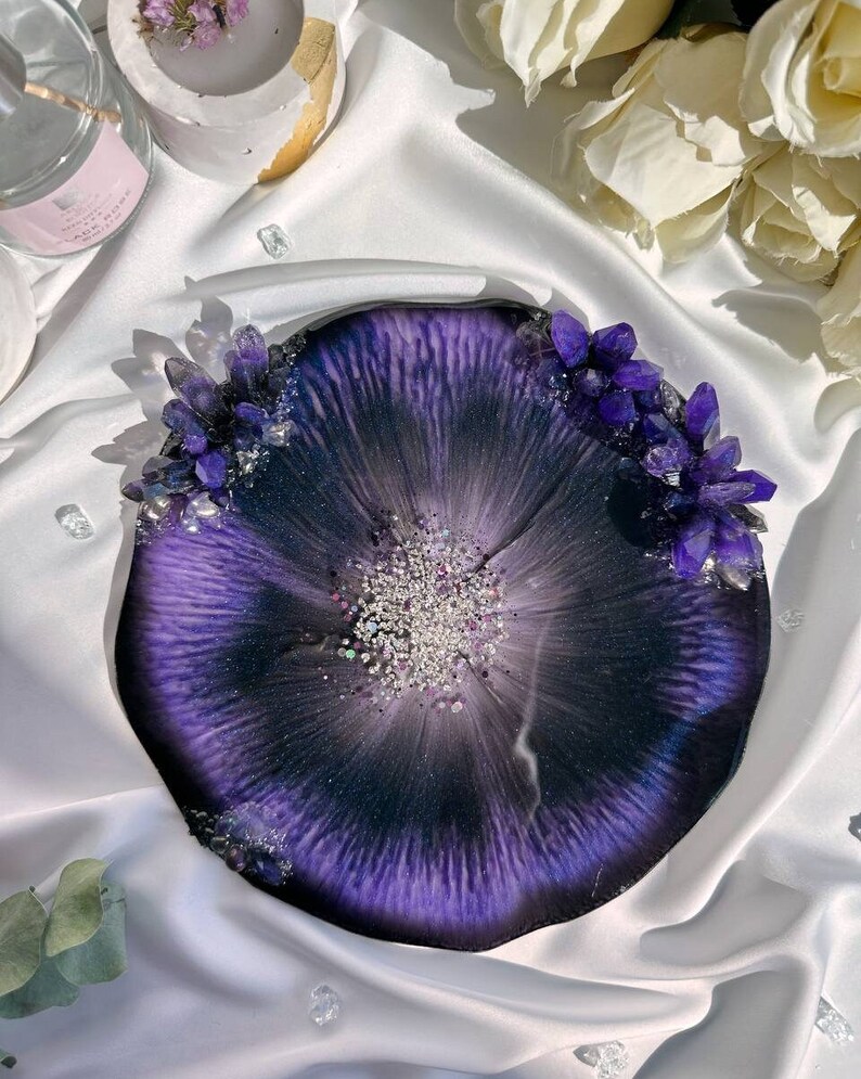 May include: A round, resin tray with a deep purple and black design, accented with silver glitter and clusters of purple crystals. The tray has a unique, organic shape and a glossy finish, perfect for home decor.