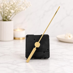 Puede incluir: Un mecanismo de reloj negro con manecillas doradas. La manecilla de la hora es un rectángulo plano, y la manecilla de los minutos es una varilla larga y delgada. El mecanismo del reloj es cuadrado y se encuentra sobre una superficie de mármol blanco.