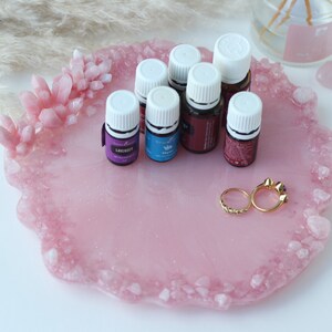 May include: A pink resin tray with a crystal-like border, holding essential oil bottles and two gold rings. The tray has a smooth surface with embedded glitter. Several small bottles with white caps are arranged on the tray.