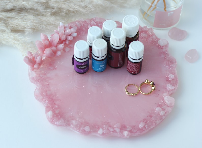 May include: A pink resin tray with a crystal-like border, holding essential oil bottles and two gold rings. The tray has a smooth surface with embedded glitter. Several small bottles with white caps are arranged on the tray.