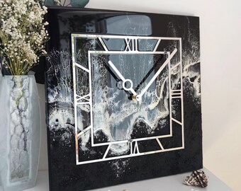 May include: Square black wall clock with a silver frame and white hands. The clock face features an abstract design in white and grey. The clock is displayed on a white surface with decorative items.