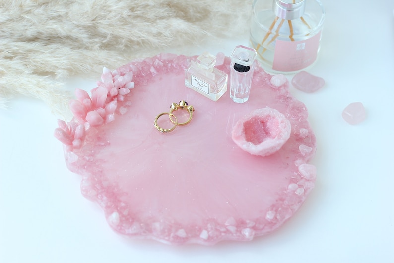 May include: A pink resin tray with a crystal-like border and accents. The tray holds gold rings, perfume bottles, and a geode. Decorative items are arranged on a white surface, creating a stylish display.