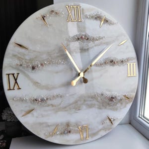 Könnte beinhalten: Eine runde Wanduhr mit einem weißen und beigefarbenen Marmormuster. Die Uhr hat goldene römische Ziffern und goldene Zeiger. Dekorative Steine sind in das Design eingebettet. Die Uhr ist ein dekorativer Einrichtungsgegenstand.