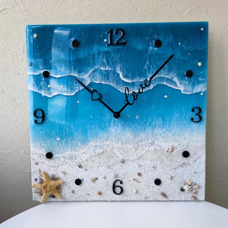 May include: A square resin clock with a blue and white ocean scene. The clock face has black hands and black numbers. The word "love" is written on the clock face with a heart symbol. The clock is surrounded by white sand and seashells.