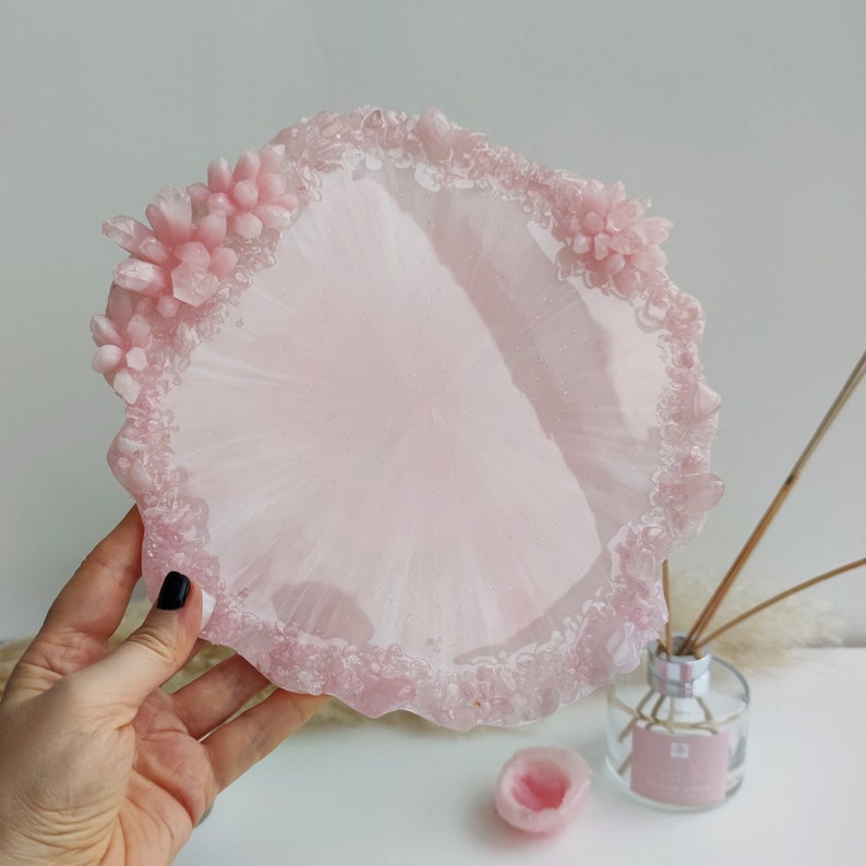 May include: A light pink resin tray with a textured, crystal-like edge. The tray has a smooth, glossy surface and is decorated with small, clustered crystal formations. The tray is round and appears to be handmade.