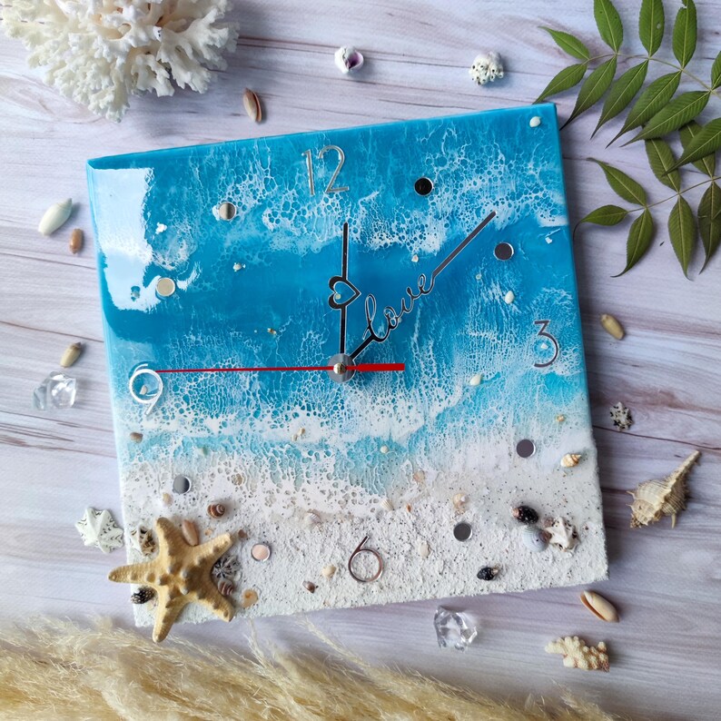 May include: Square resin clock with a blue and white ocean wave design. The clock face has white numbers and a red hour hand and black minute hand. The clock has a white sand beach design at the bottom with seashells and a starfish. The clock face also has the word "Love" written in cursive with a heart.