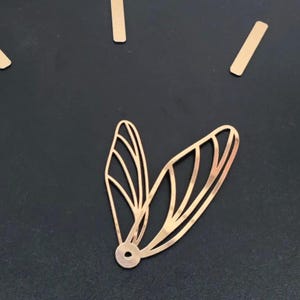 Può includere: Un set di delicati ciondoli in metallo color oro a forma di ali di libellula e diversi pezzi rettangolari, forse per la creazione di gioielli o l'artigianato. Le ali hanno intricati dettagli a forma di venature e una base circolare.