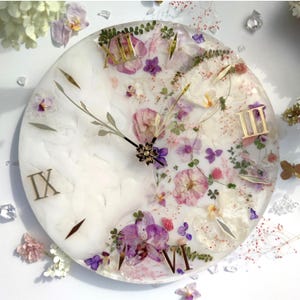 May include: A white round clock face with Roman numerals and gold accents. The clock face is covered in pressed flowers and greenery, creating a floral design.