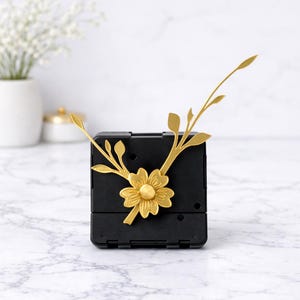May include: A black square clock mechanism featuring decorative gold-coloured clock hands shaped like a flower with branching leaves. The central flower element serves as the hub for the two slender, leaf-adorned hands extending outward.
