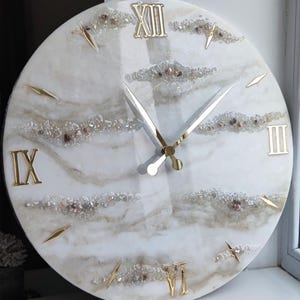 May include: A round wall clock with a white and beige marble-like design. The clock features gold Roman numerals, gold hands, and gold markers. The clock face is decorated with small, sparkling crystals.