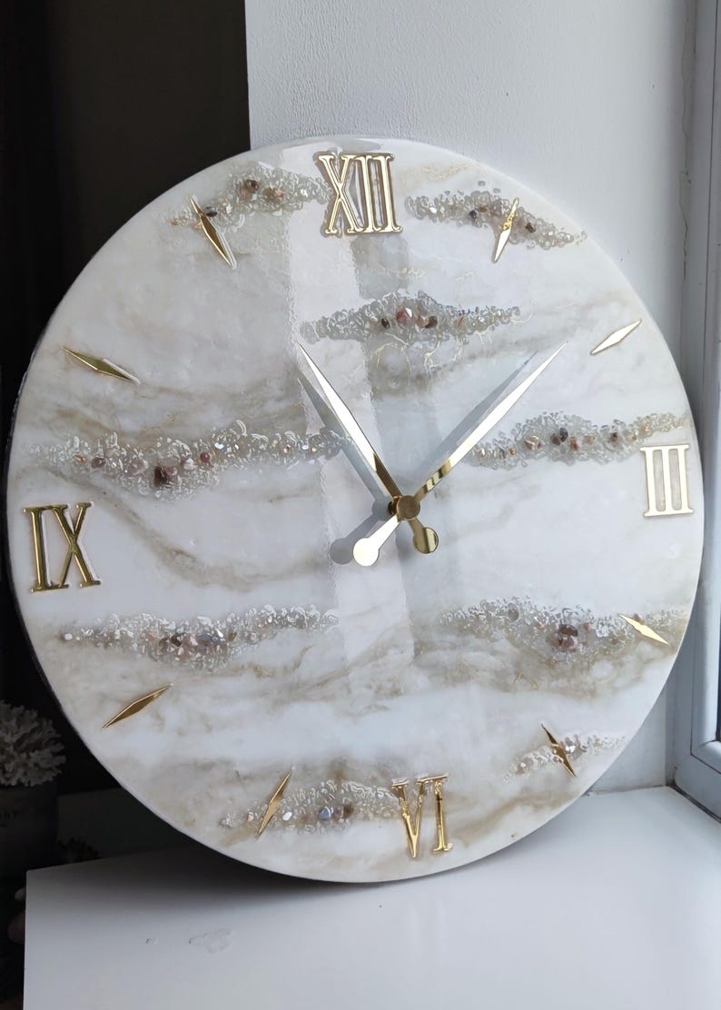May include: A round wall clock with a white and beige marble-like design. The clock features gold Roman numerals, gold hands, and gold markers. The clock face is decorated with small, sparkling crystals.