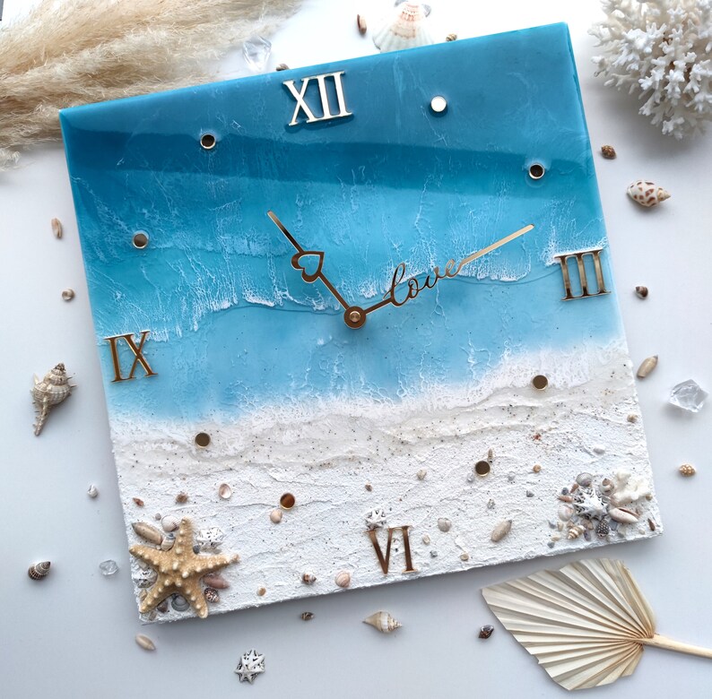 May include: A square resin clock with a blue and white ocean scene. The clock face has Roman numerals and the word "love" written in cursive. The clock is surrounded by white sand and seashells.