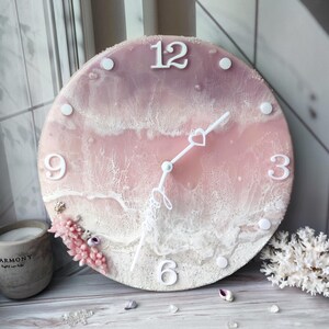 Peut inclure: Une horloge murale ronde avec un design sur le th&egrave;me de l'oc&eacute;an rose et blanc. Le cadran de l'horloge comporte des chiffres et des aiguilles blancs, avec un d&eacute;tail en forme de c&oelig;ur sur l'aiguille des secondes. Des &eacute;l&eacute;ments d&eacute;coratifs incluent du corail et des coquillages.