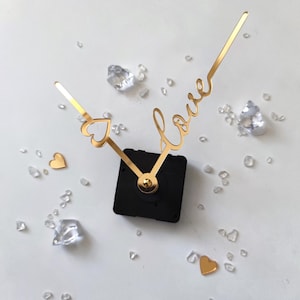May include: Gold clock hands with a heart and the word "love" written in cursive. The hands are attached to a black clock mechanism.