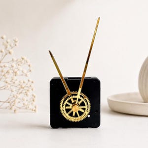 May include: A black square clock movement mechanism featuring a gold-coloured gear-shaped centre and two long, slender gold-coloured clock hands extending upwards. The mechanism is designed for timekeeping and displays a metallic finish on the hands.