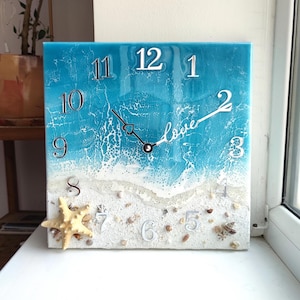 May include: A square resin clock with a beach scene. The clock face is blue with white waves and a sandy beach with seashells. The numbers are white and the hands are black. The words "love" are written on the clock face.