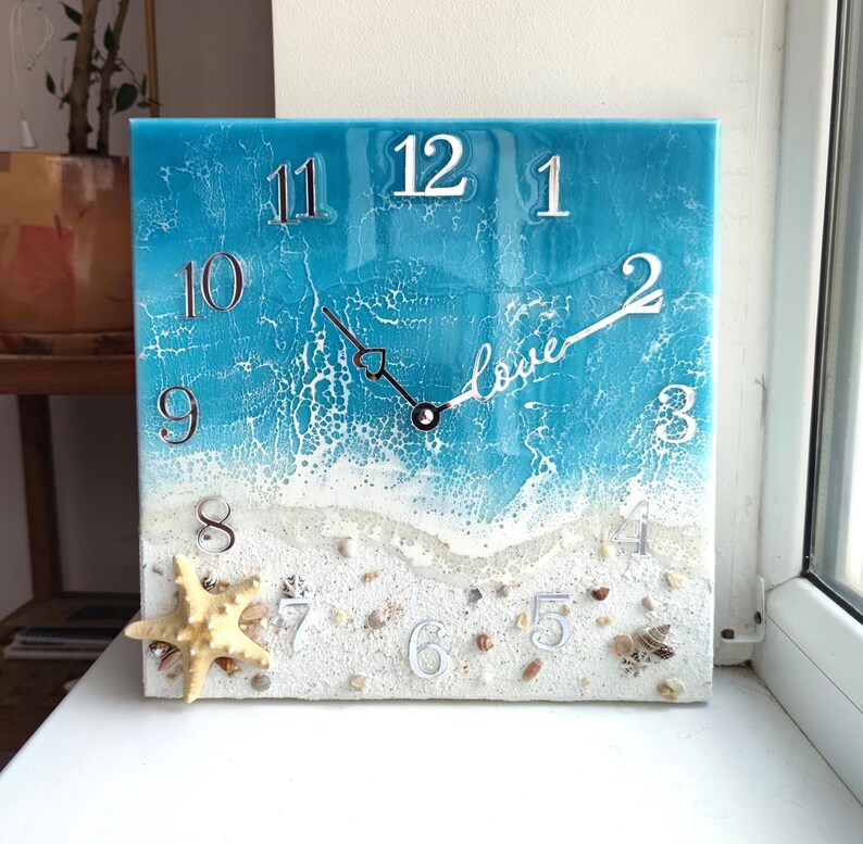 May include: A square resin clock with a beach scene. The clock face is blue with white waves and a sandy beach with seashells. The numbers are white and the hands are black. The words "love" are written on the clock face.
