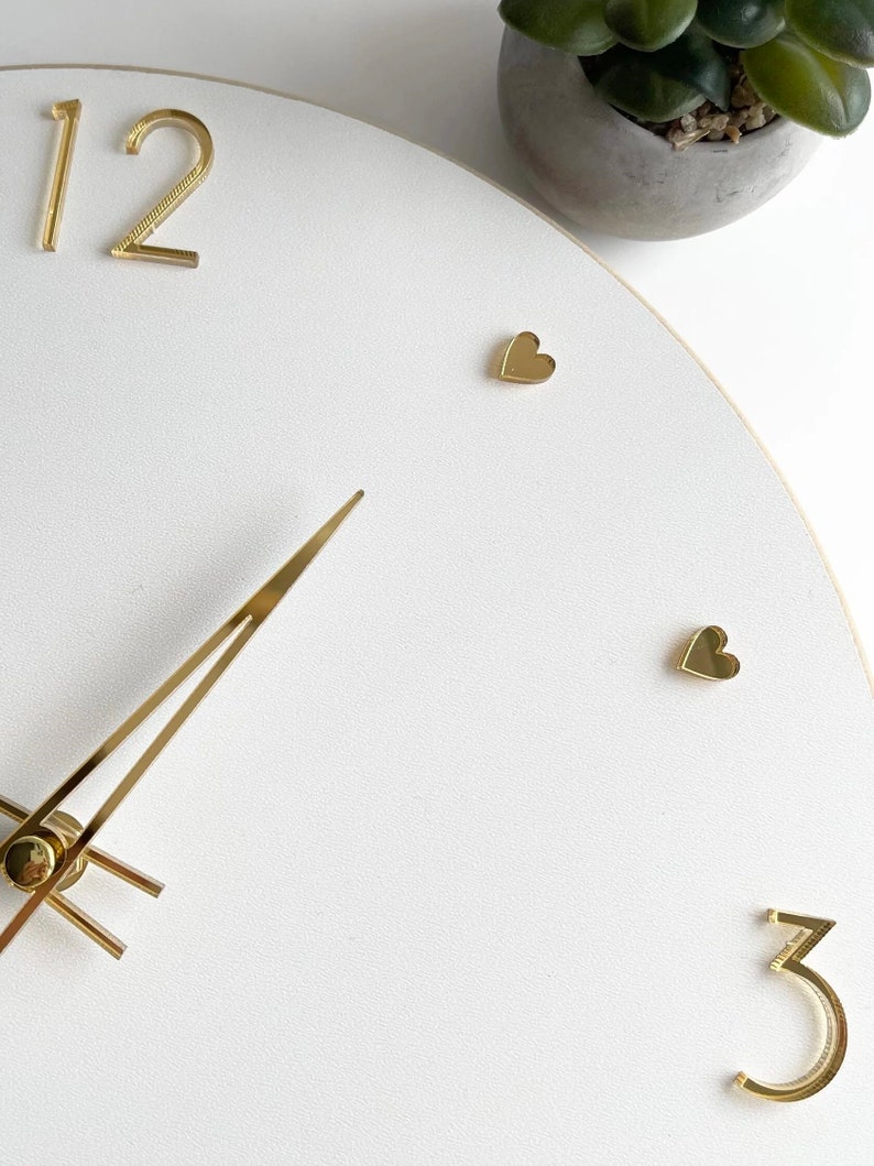 Peut inclure: Horloge murale blanche avec des aiguilles et des chiffres dor&eacute;s. Le cadran comporte des marqueurs en forme de c&oelig;ur dor&eacute;s et les chiffres 12 et 3. Une petite plante en pot est visible en arri&egrave;re-plan.