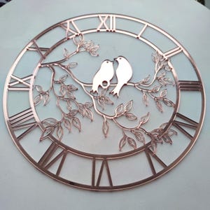 May include: A decorative wall clock with a rose gold finish. The clock face features Roman numerals and a branch with two birds. The birds are perched on a branch with leaves, adding a touch of nature to the design.