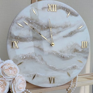 May include: A round wall clock with a white and gold design. The clock face features gold Roman numerals and hands, with a cloudy, marbled effect. The clock is displayed on a wooden easel, with a string of pearls and a bouquet of roses.