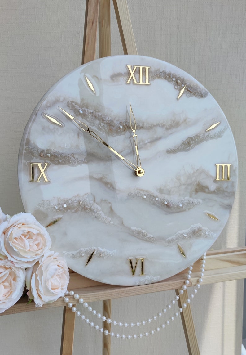 May include: A round wall clock with a white and gold design. The clock face features gold Roman numerals and hands, with a cloudy, marbled effect. The clock is displayed on a wooden easel, with a string of pearls and a bouquet of roses.