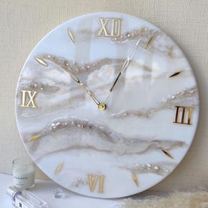 May include: A round wall clock with a white and beige marbled design. Gold Roman numerals mark the hours, and gold hands indicate the time. The clock has a diameter of approximately 12 inches.