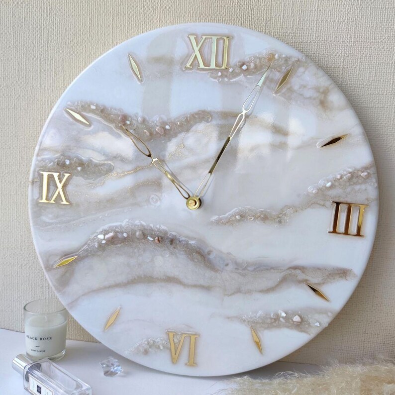 May include: A round wall clock with a white and beige marbled design. Gold Roman numerals mark the hours, and gold hands indicate the time. The clock has a diameter of approximately 12 inches.