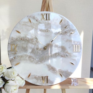 May include: Round wall clock with a white and beige marbled design. The clock features gold Roman numerals and gold hands. The clock's face has a textured, cloud-like appearance with embedded decorative elements.