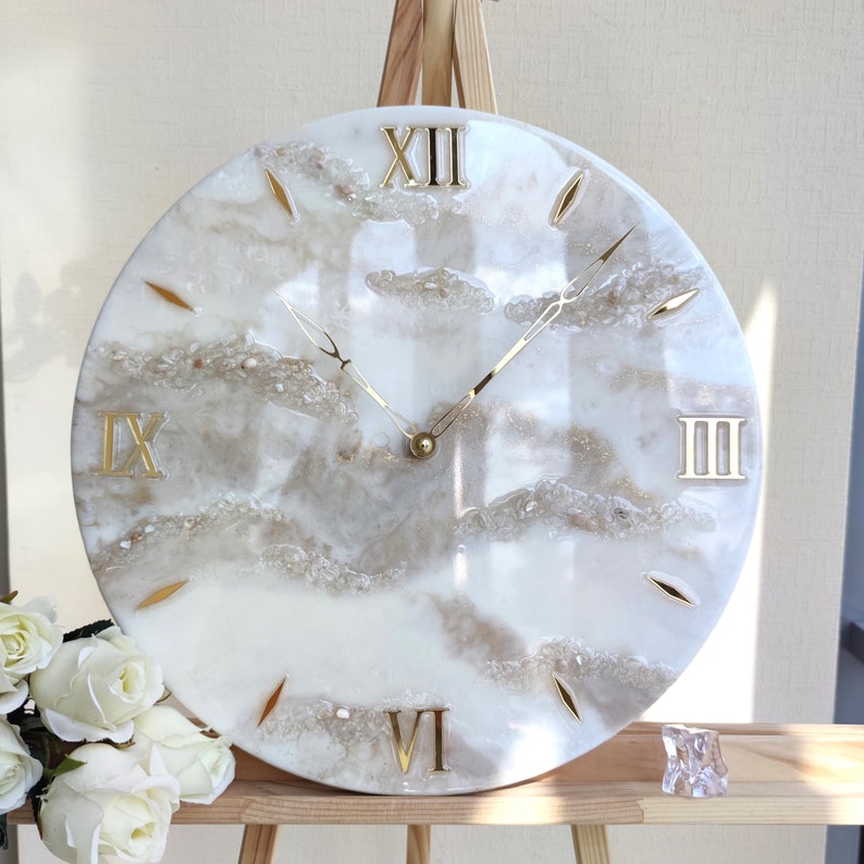 May include: Round wall clock with a white and beige marbled design. The clock features gold Roman numerals and gold hands. The clock's face has a textured, cloud-like appearance with embedded decorative elements.