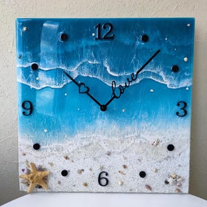 May include: A square resin clock featuring a blue and white ocean wave design. The clock face has black numbers and hands, and the word "love" is written in cursive on the face. The clock is surrounded by white sand and seashells.