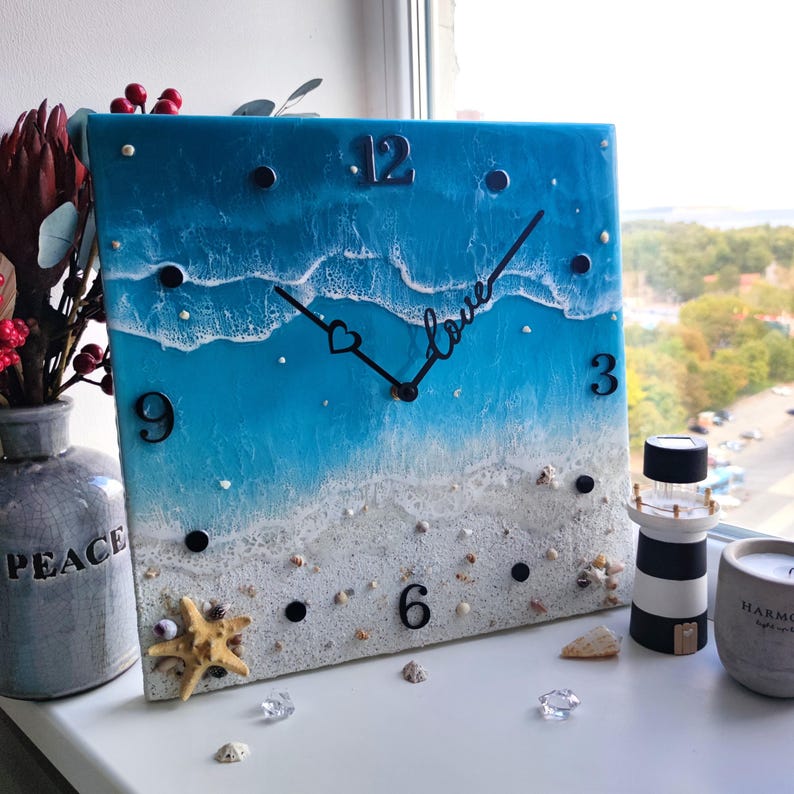 Peut inclure: Horloge murale carr&eacute;e avec un motif de plage. Le cadran de l'horloge pr&eacute;sente une sc&egrave;ne d'oc&eacute;an bleu et blanc avec des coquillages et une &eacute;toile de mer. Les aiguilles noires indiquent l'heure, et le mot "love" est &eacute;crit en cursive.