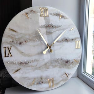 May include: A round wall clock with a white and beige marbled face. Gold Roman numerals mark the hours, and gold hands indicate the time. The clock face is decorated with small, sparkling crystals and stones.
