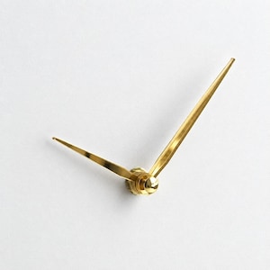 May include: A pair of polished gold-tone clock hands featuring a sleek, tapered design. The two hands are connected at a central circular hub, with one hand significantly longer than the other, both extending outward from the centre point.