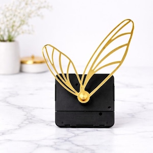 May include: A black clock movement with gold-colored butterfly wings. The clock mechanism has a square shape and a gold-colored center. The wings are open and have a delicate, airy design. The background is a marble surface.