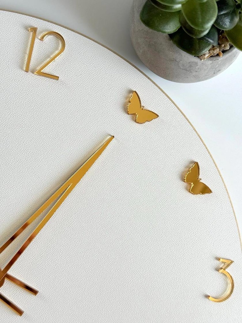 Peut inclure: Une horloge murale blanche avec des aiguilles, des chiffres et des papillons dor&eacute;s. L'horloge affiche les chiffres 12 et 3, avec deux papillons d&eacute;coratifs. L'horloge est un objet d&eacute;coratif.