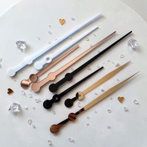 May include: A collection of clock hands in various colors, including white, rose gold, black, and gold. The hands are arranged on a white surface with decorative crystals and gold heart-shaped accents.