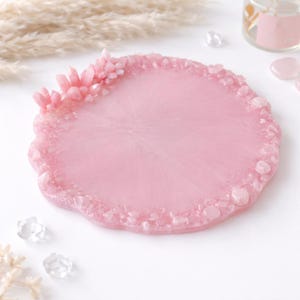 May include: A round, light pink resin coaster with a textured edge and embedded crystals. A cluster of pink crystals adorns one side. The coaster is a decorative item, perfect for holding a glass or mug.