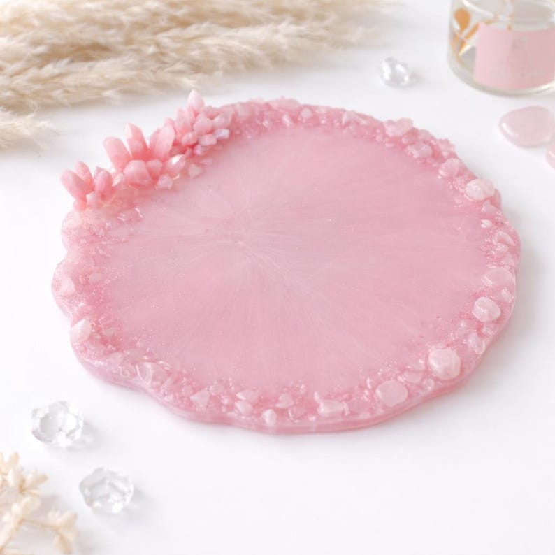 May include: A round, light pink resin coaster with a textured edge and embedded crystals. A cluster of pink crystals adorns one side. The coaster is a decorative item, perfect for holding a glass or mug.