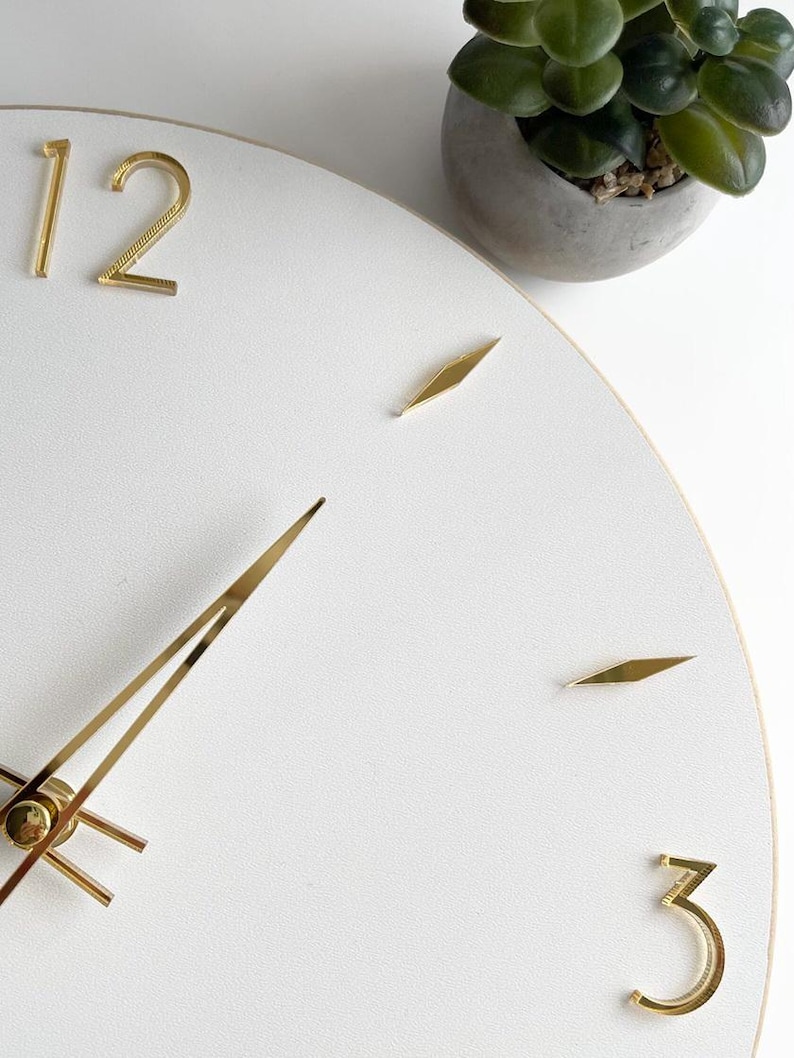Peut inclure: Horloge murale blanche avec des aiguilles et des chiffres dor&eacute;s aux positions 12 et 3. La montre a une bordure dor&eacute;e et est &agrave; c&ocirc;t&eacute; d'une petite plante en pot. Diam&egrave;tre d'environ 30 cm.