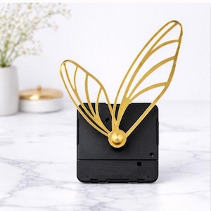 Könnte beinhalten: Ein Uhrwerk mit zwei goldfarbenen Metallzeigern in Form von zarten Insektenflügeln. Die Zeiger sind an einem quadratischen schwarzen Kunststoffgehäuse mit einer mittigen, goldfarbenen runden Kappe befestigt, das zur Wandmontage gedacht ist.