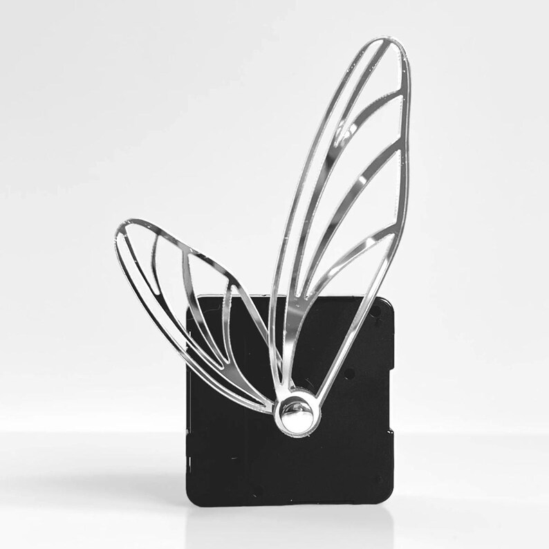 May include: A silver-colored, butterfly wing-shaped decorative object attached to a black square base. The wing design features multiple curved lines, creating an open, airy appearance. The base has a small, circular silver accent.