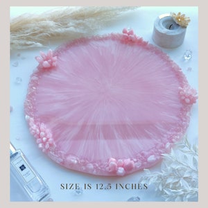May include: A round, pink resin tray with a crystal-like border and decorative crystal clusters. The tray is 12.5 inches in diameter. The surface has a swirled, translucent design. The text "SIZE IS 12.5 INCHES" is at the bottom.