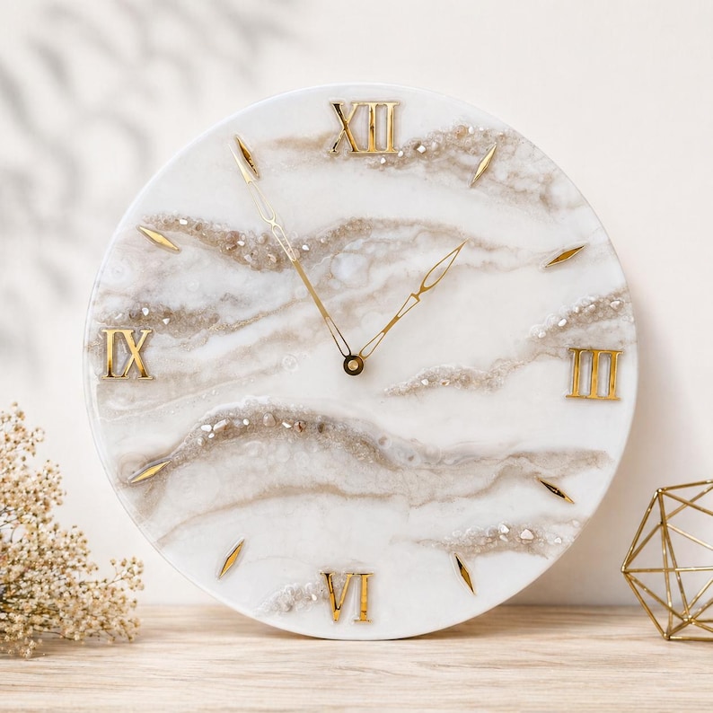 May include: A round wall clock with a white background and a marbled, beige design. The clock features gold Roman numerals and gold hands. The clock is approximately 12 inches in diameter.