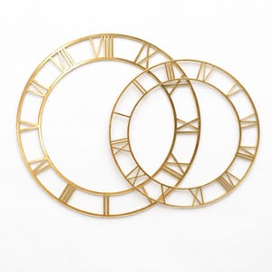 Peut inclure: Deux cadrans d'horloge en métal doré avec des chiffres romains. Le plus grand cadran est positionné derrière le plus petit, créant un design superposé. Les cadrans sont posés sur un fond blanc.