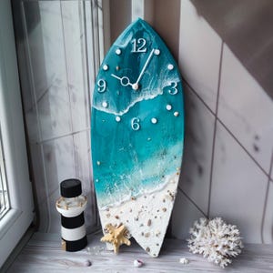 Peut inclure: Horloge murale en forme de planche de surf avec un design sur le th&egrave;me de la plage. Le cadran de l'horloge pr&eacute;sente une sc&egrave;ne d'oc&eacute;an turquoise avec des vagues blanches, des coquillages et une &eacute;toile de mer. Des chiffres blancs indiquent les heures.